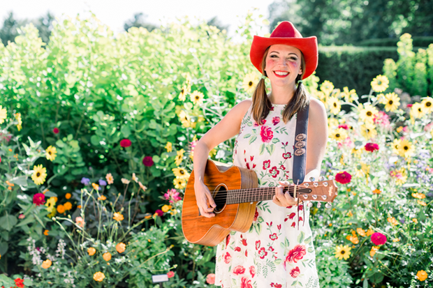 Woman in a flowered dress playing a guitar wearing a red cowboy hat.
