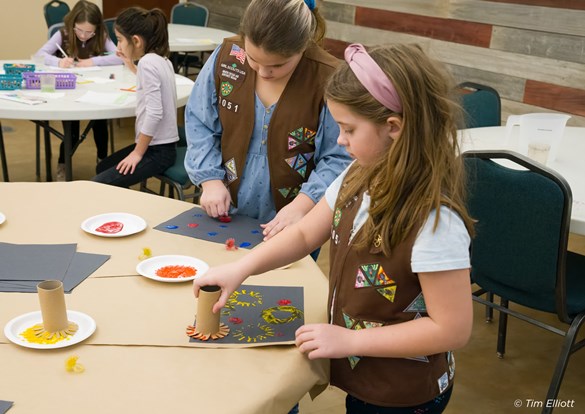 Two young girls in the Girl Scout uniforms doing arts and crafts.