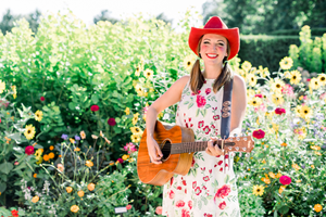 Woman in a flowered dress playing a guitar wearing a red cowboy hat.