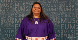 Native American woman dressed in a purple dress with red and white belt.