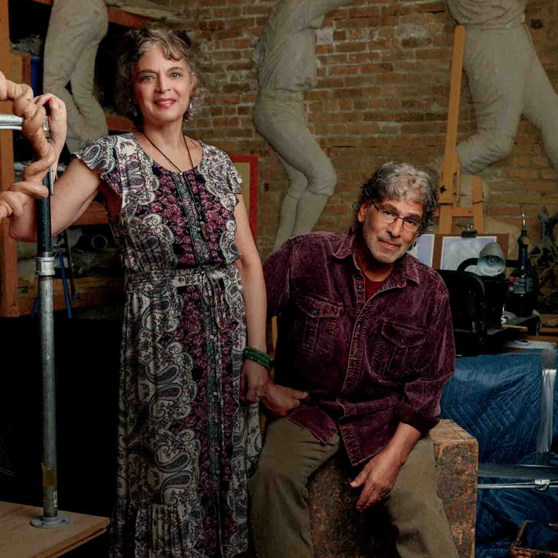 Woman standing in flowered dress and a man sitting next to in front of large sculptures