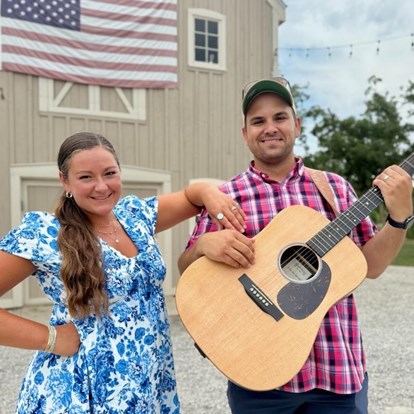 Woman in blue/white flowered dress and man in red plaid shirt holding a guitar.