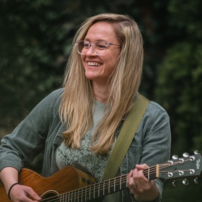 Woman with long blonde hair wearing glasses, playing a guitar.