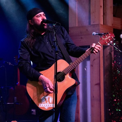 Man in dark shirt with a black hat, playing a guitar on stage.