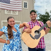 Woman in blue/white flowered dress and man in red plaid shirt holding a guitar.