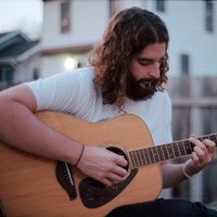 Man with long hair and beard, wearing white t-shirt playing a guitar.