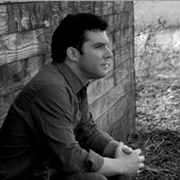 Man with dark hair in a dark shirt leaning against a barn.