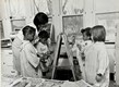 1950's Black female teacher helping black and white children paint on an easel.