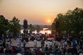 A diverse crowd stands around a stage, with a beautiful sunset creating a colorful backdrop.