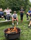 Young men and woman sitting on picnic tables in the woods, enjoying the fire pit.