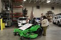 Group of people in a garage gathered around a green lawnmower.