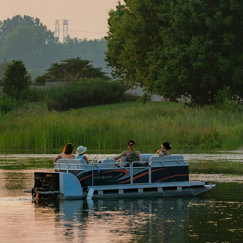 A pontoon boat floating on a tranquil lake with gentle ripples, bordered by trees and a clear blue sky above