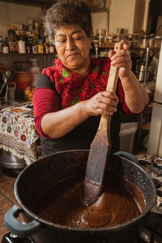 Hispanic woman in a red rose dress, stirring a large pot with a wooden paddle.