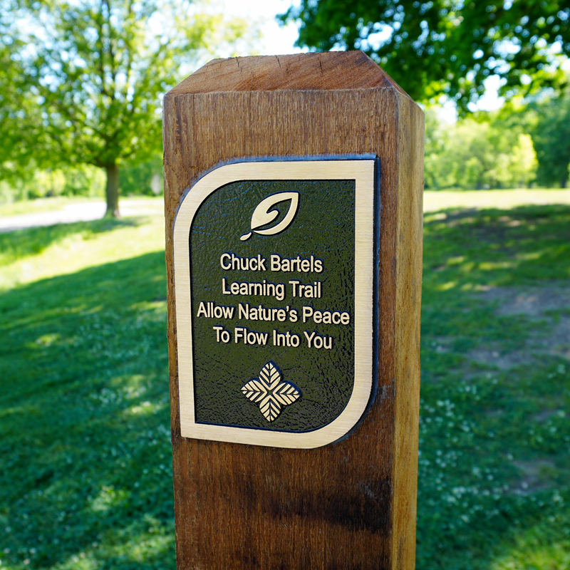 Wooden post with a brass plaque that read "Chuck Bartels Learning Trail Allow Natures Peace To Flow Into You"