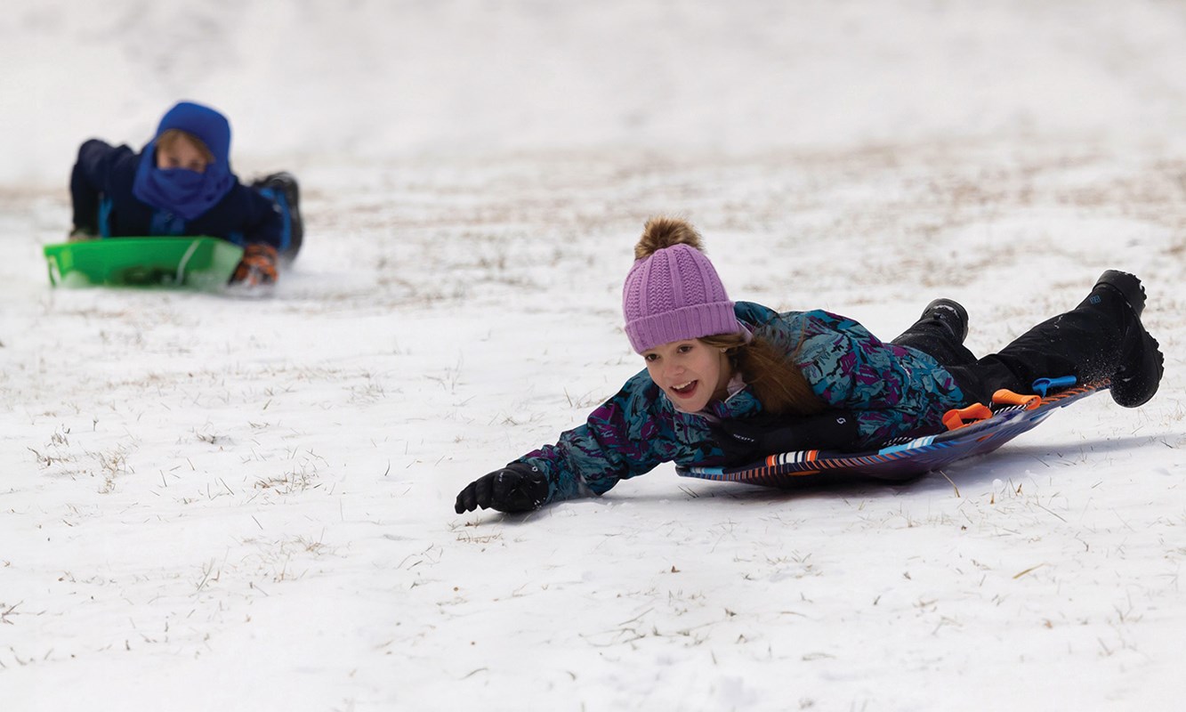 Two kids sledding down a hill on separate sleds, both on their bellies.