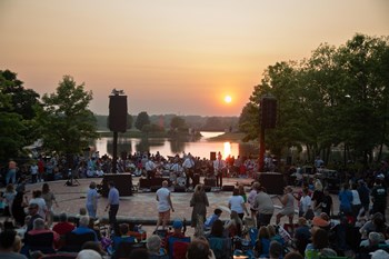 A diverse crowd stands around a stage, with a beautiful sunset creating a colorful backdrop.