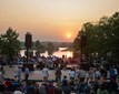 A diverse crowd stands around a stage, with a beautiful sunset creating a colorful backdrop.